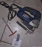 Full view of blue Ryobi electric laser jigsaw with attached power cord, red-handled reamer tool next to it.