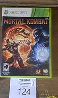 Photo of Xbox 360 Mortal Kombat game case with visible cover art showing characters Scorpion and Sub-Zero, green game case edge, and auction lot tag 124.