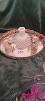 Photo showing an old oval mirror tray with metal decorative rim, a hobnail style white glass decanter, and two small teddy bears: one with a red ribbon and one with a striped scarf. A small heart decoration is also visible on the mirror.