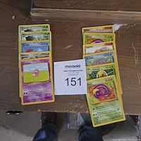 Photo of 17 Pokémon cards arranged in two columns on a wooden surface with a MaxSold auction tag marked 151.