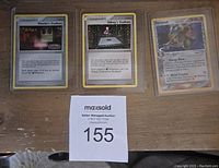 Three Pokemon cards in protective sleeves laid out on a wooden surface, two Trainer cards (Phoebe's Stadium, Sidney's Stadium) and one Metal type Marshtank card.
