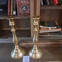 Two solid brass candle holders shown together on a table with bookshelf background, displaying size and design