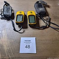 Two yellow Garmin eTrex handheld GPS units with connected wrist straps and two protective cases on wooden surface with MaxSold lot card 48.