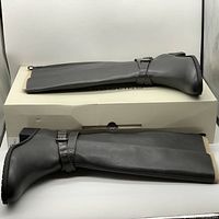 Side view of the pair of dark gray Bandolino Noles tall shaft women's boots with buckle strap detail, lying on top of original box.
