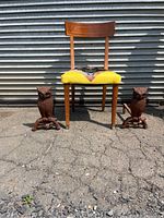 Photo of wooden chair with yellow upholstered seat flanked by two cast iron owl andirons placed on pavement in front of metal door.