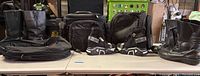 Wide angle shot showing two pairs of black leather motorcycle boots, black and white riding shoes, black motorcycle bags on a table.
