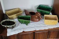 Front view of six vintage ceramic planters including a duck planter, two rectangular green and yellow planters, and three other shapes on a white cloth over cabinet.