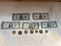 Top-down photo showing five US paper bills and a row of nine US coins beneath on a beige surface.