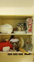 Photo of baking and kitchen items inside cupboard including a large white insulated hot and cold dish with lid on top shelf and various glass and metal baking dishes, a box with a metal spring form pan, and package-wrapped items.