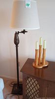 Floor lamp standing next to a wooden cabinet with three-stick wooden candle holder on top, showing lamp base, shade, and candle holder