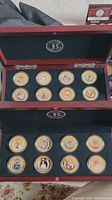 Wide shot showing two rows of eight 24K gold plated Princess Diana coins in a wooden display case with fabric lining and embossed branding.