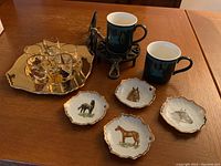 Photo showing coffee warmer with horse head, two dark coffee cups, six shot glasses on gold plate, and four small horse decorated plates.