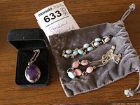 Photo showing amethyst pendant in a black jewelry box, silver chain necklace, and a grey pouch holding a bracelet with turquoise stones and matching necklace and bracelet with pink and blue beads.