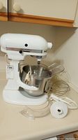 White Kitchen Aid stand mixer model K5SS on counter with stainless steel mixing bowl attached, splatter guard, and whisk attachment visible.