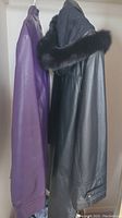 Side view of black leather coat with fur hood and purple leather-like jacket hanging on rack.