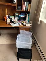 Three clear Staples plastic letter file boxes stacked, with a black laptop bag in front, on carpet beside a wooden desk filled with various office supplies.