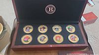 Full set of 8 gold-plated Queen Elizabeth II coins in burgundy wooden display case with black padded interior.
