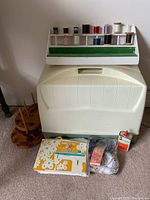Singer sewing machine and accessories with thread spools on wooden riser, oil can, and instruction booklet in front of portable carrying case