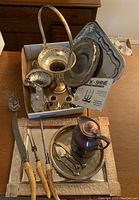 Box containing various silver plated items including trays, candlesticks, salt and pepper shakers, and a silver pitcher, with carving set laid out in front.