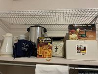 Shelf showing a variety of small kitchen appliances and utensils including an apple parer, corer and slicer in box, kitchen scale, cookie press, pasta pot box, coffee urn with cross stitched cover and carafes.