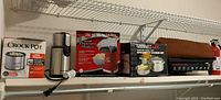 Full lot view on shelf showing mini Crock-Pot Little Dipper, Betty Crocker popcorn maker, Gourmet Magic spaghetti cooker set, KitchenAid grinder, Charles Craft grill in bag.