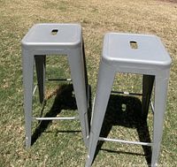 Two gray metal bar stools shown outside on grass, with square tops and cut-out handles in the center.