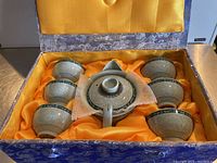 Full tea set in decorative blue box with yellow satin lining, showing six cups around central teapot with foam protector.