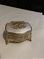 Closed view of the small ornate metal jewelry box showing detailed floral and leaf embossing and claw feet.