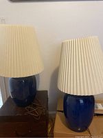 Full view of two blue ceramic base table lamps with pleated cream-colored shades placed on dark and cardboard boxes against a white wall.