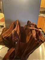 Front and side view of well worn brown leather attaché case showing multiple zippered compartments and carrying handles.