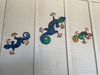 Four metal geckos hanging on a white wall showing size and color variations