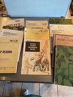 Eleven military field manuals arranged side by side. Includes titles on map reading, chemical accident contamination, NATO air force, military leadership, and chemical operations.