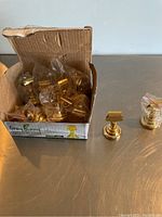 Box containing multiple gold-colored name card holders in plastic bags and two individual holders placed on surface