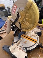Close-up of Ryobi 10 inch blade with yellow transparent plastic guard and Ryobi logo visible. Shows part of the base and dust bag.