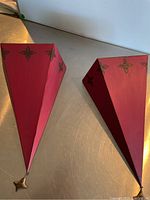 Photo showing two red metal hanging shelves with pointed bottom design and decorative four-petal motifs on top edges, placed on a silver surface.