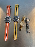 Photo of four wristwatches on a metallic surface: two Fossil watches with fabric straps (orange and yellow), one gold-tone Fossil metal bracelet watch, and one silver-tone Disney Mickey Mouse watch with metal bracelet.