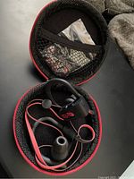 Open carrying case showing black and red in-ear headphones with ear hooks and mesh pocket containing small accessories.