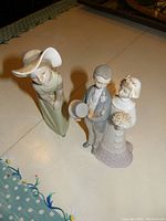 Two Lladro porcelain figurines on display surface, one is a woman in a green dress and hat, the other is a bride and groom couple.