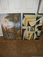 Photo of two unframed canvas oil paintings standing against a white door, showing the forest landscape painting and the abstract geometric painting side by side on the floor.