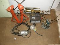 Photo showing two orange metal jack stands, handheld electric polisher or sander with cord, blue hydraulic bottle jack, handheld work light, and metal toolbox on floor.