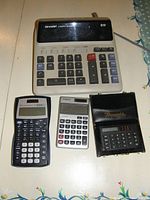 Photo showing four calculators: one large Sharp desktop calculator, one Casio scientific calculator, one Casio basic calculator, and one Pequeño compact calculator with cover.