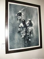 Framed black and white print photograph showing three dirt bike riders in racing action, with a dark frame and white matting.