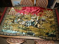 Wide shot of the full tapestry laid out on a table showing two Arabian horsemen riding horses with palm trees and a building in the background.