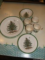 Place setting plates, bowls, and cups grouped on a table showing the Christmas tree pattern and green rim detail.