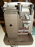 Front angled view of textured beige Bell & Howell movie projector showing control knobs, lens, and side compartment.