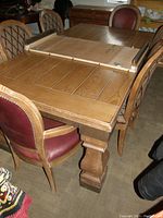 Photo showing the wooden dining table with one leaf, rectangular shape, natural finish and detailed pedestal base, surrounded by chairs.