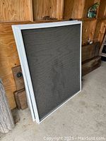 Photo showing a stack of large square aluminum framed screens leaning against a wooden wall.
