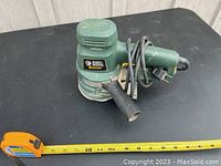 Green Black & Decker Quantum electric orbital sander with handle and power cord, placed next to a measuring tape showing approximately 11 inch length.