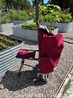 Side view of foldable burgundy nylon shopping cart with attached fold-down seat and stair climbing wheels.