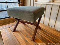 Full side view of the footstool showing greenish-blue velvet cushion and wooden X-shaped legs on a wooden surface.